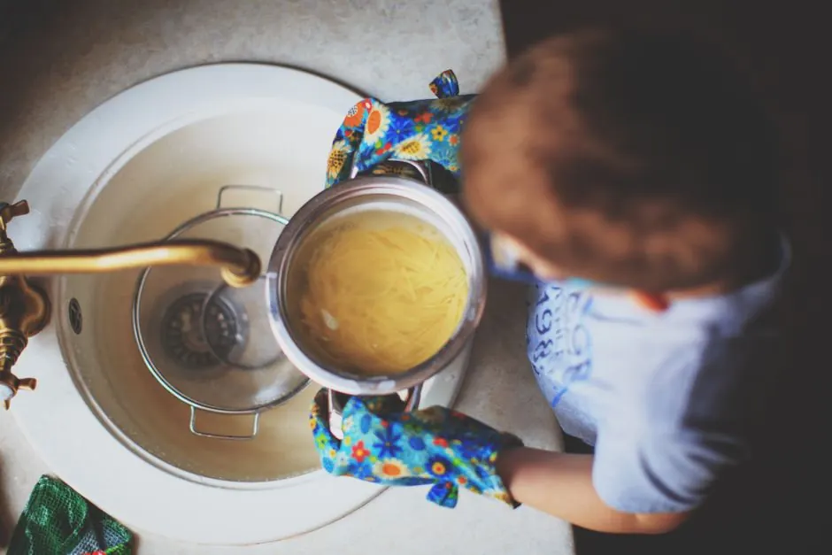 A child strains spaghetti at the sink wearing colorful oven mitts, highlighting a child's culinary exploration.